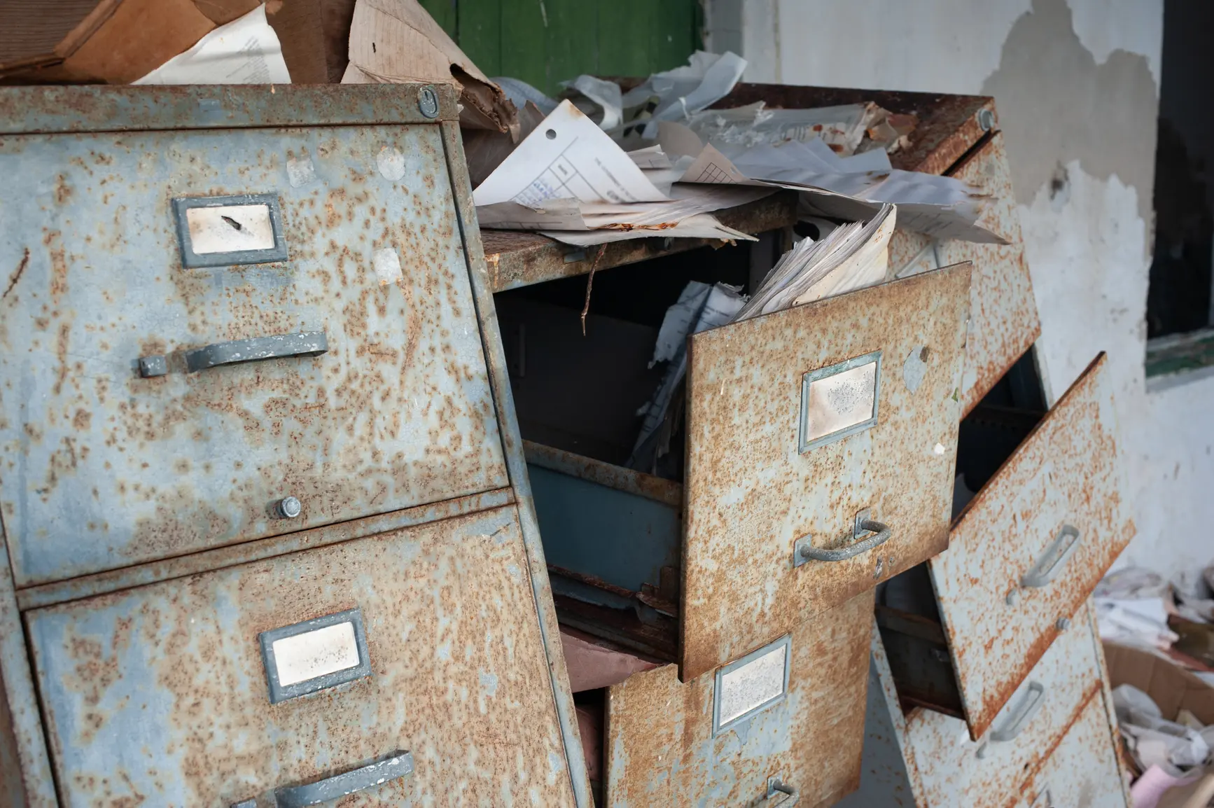 A rusty, abandoned filing cabinet with multiple drawers ajar, spilling out disorganized papers. The cabinet shows signs of significant wear and corrosion, with documents scattered on top and around it, showing signs of age and use.