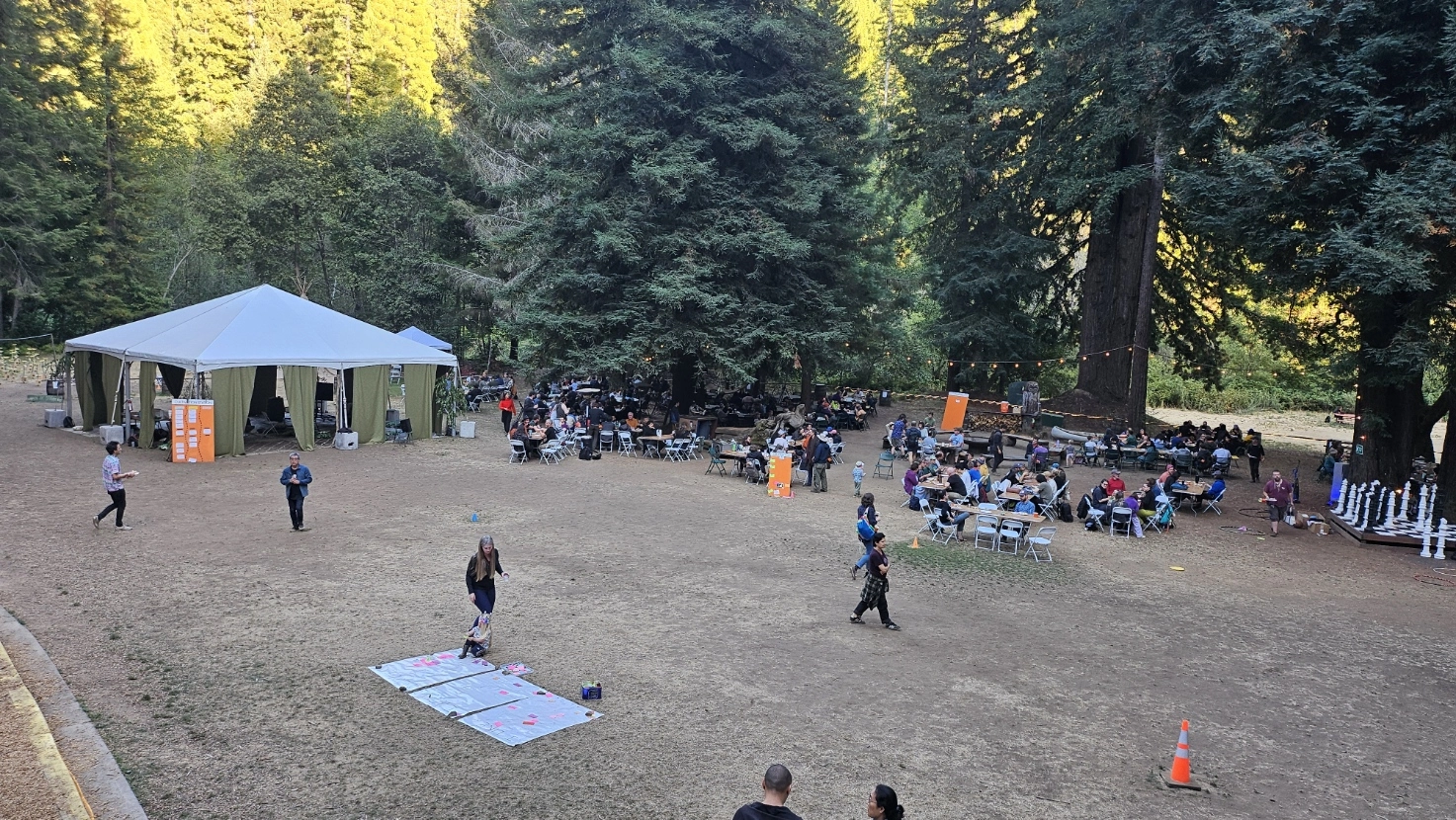 An outdoor gathering at DWeb Camp 2024 in a forested setting, surrounded by tall redwood trees. Attendees are seated at multiple tables. A large white canopy tent with open sides is set up on the left. To the right, a giant chess set is visible on a platform. Several people are walking around the open space and string lights hang between the trees.