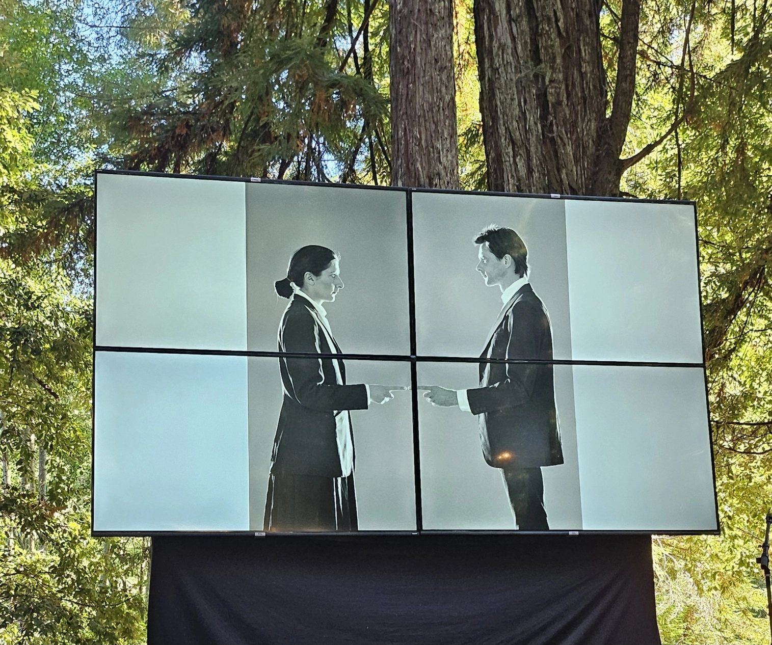 A large outdoor screen displays a black-and-white image of Marina Abramović and Ulay performing 'Point of Contact.' The image shows the two artists in formal attire facing each other, reaching out with their hands nearly touching. The screen is divided into four panels, set against a backdrop of tall trees and greenery.