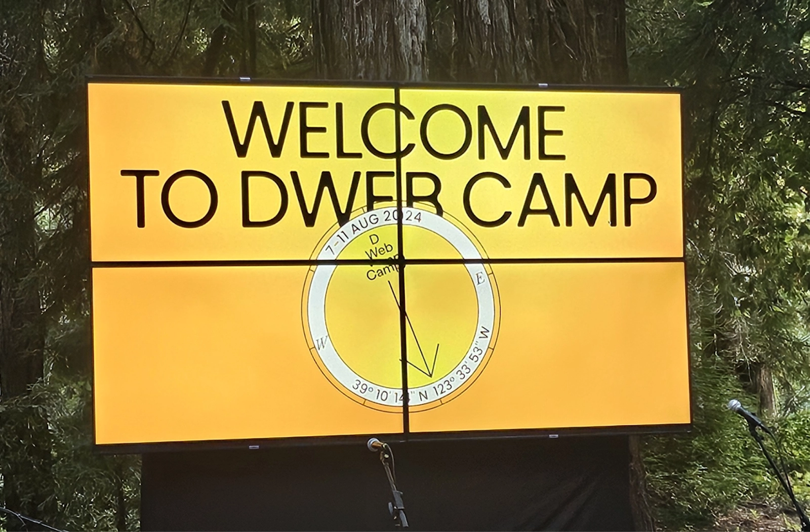 A set of 4 screens in an outdoor setting welcomes attendees to DWeb Camp 2024. The screen displays the text 'Welcome to DWeb Camp' in bold, black letters against a yellow background. Below the text is a circular compass graphic with the camp dates '7-11 AUG 2024' and the coordinates '39°10'12"N, 123°33'53"W.' The screen is surrounded by tall redwood trees.