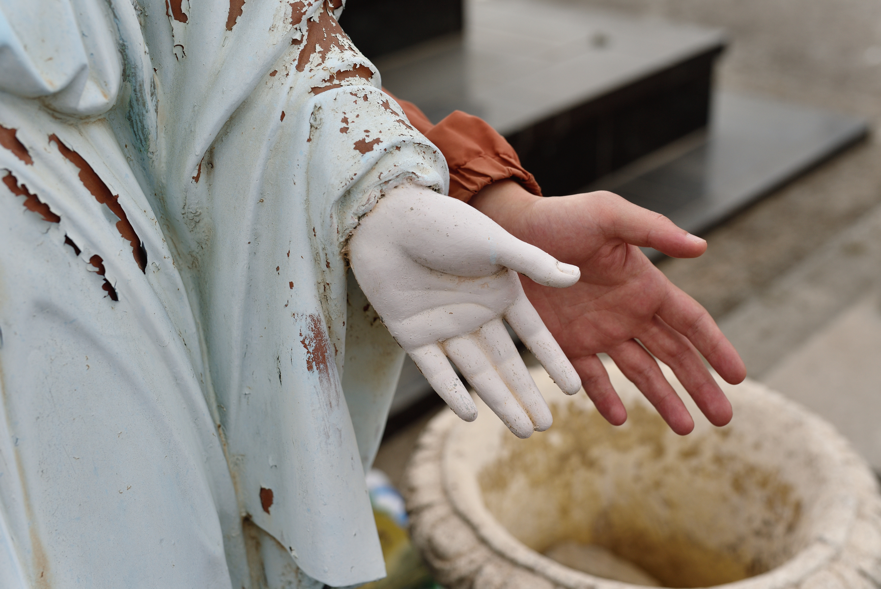A close-up of a human hand mirroring the open-palm gesture of a weathered, peeling statue. Both hands reach out over a stone basin against a blurred outdoor background.